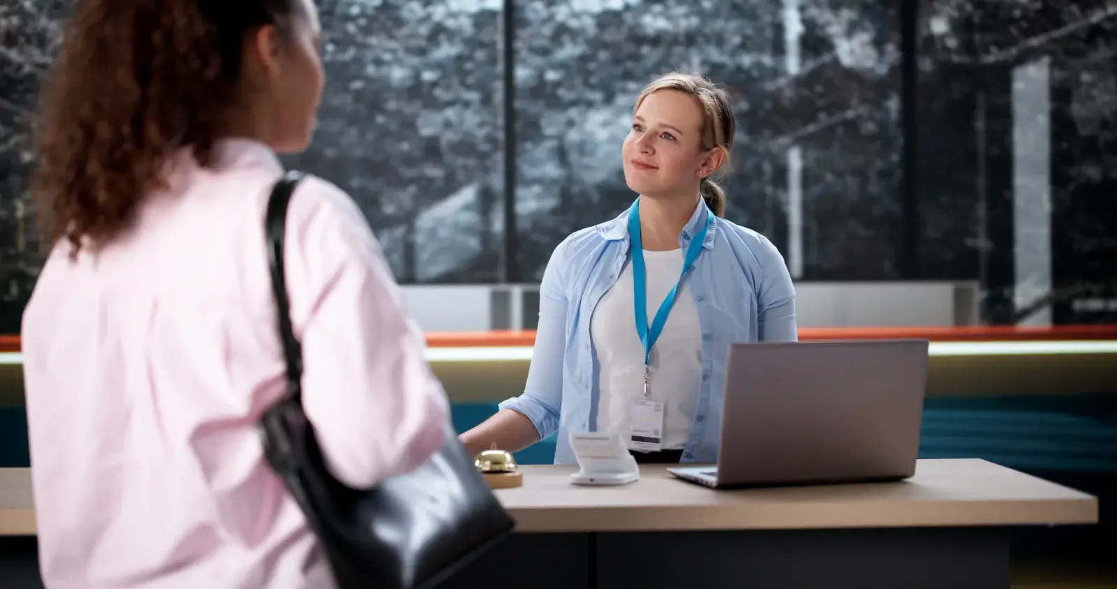 Staff member with a lanyard and laptop greets an arriving guest at a check-in desk