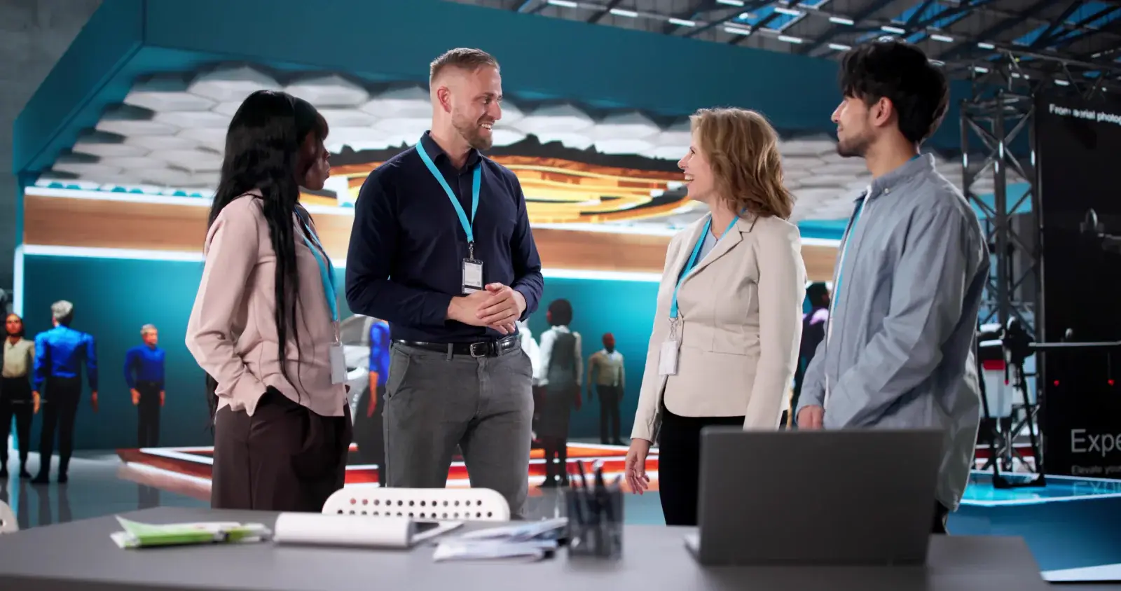 Four exhibitors wearing lanyards chat at a booth counter with a laptop on a trade show floor