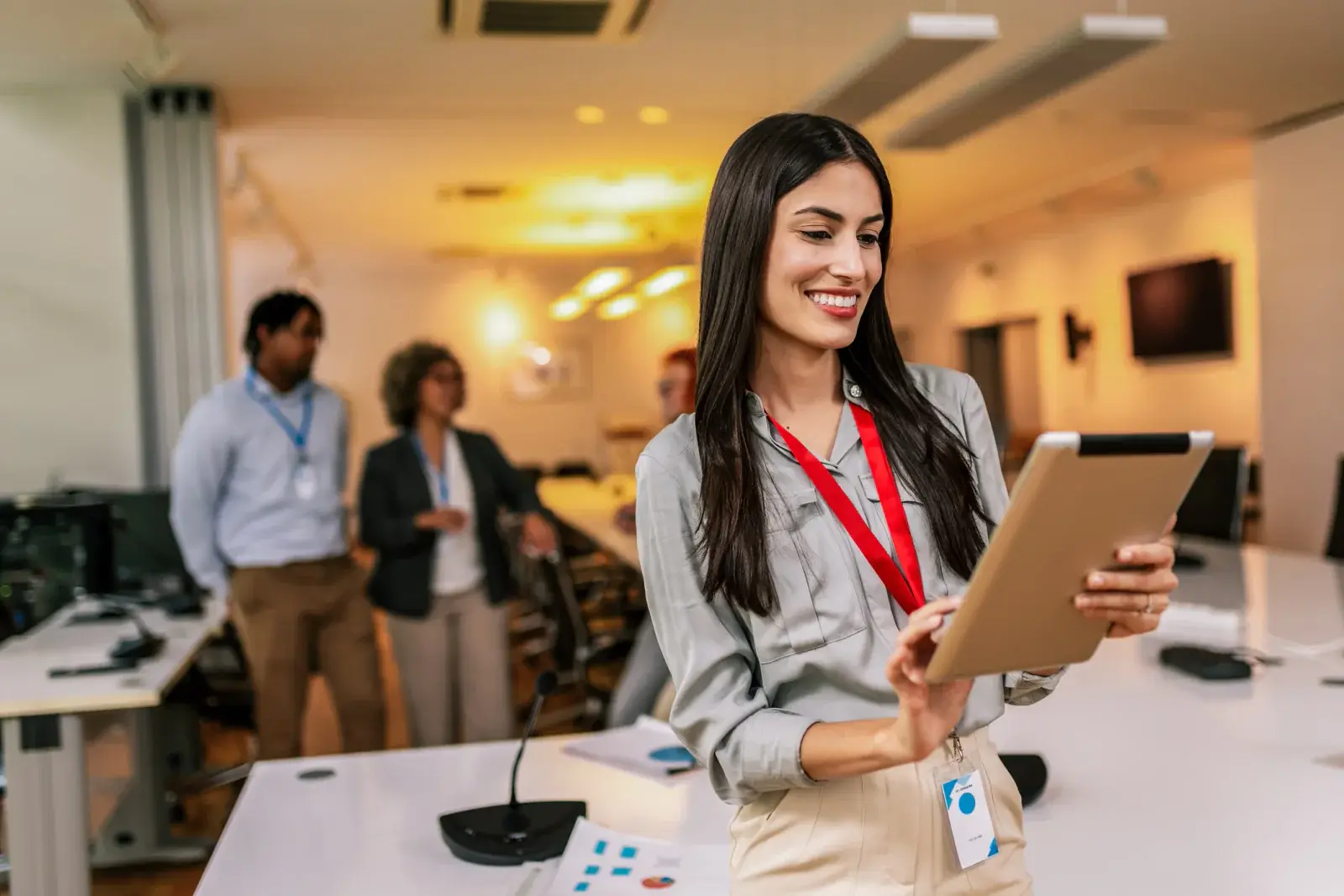 Smiling woman with a red lanyard holding a tablet in a meeting room, colleagues behind her