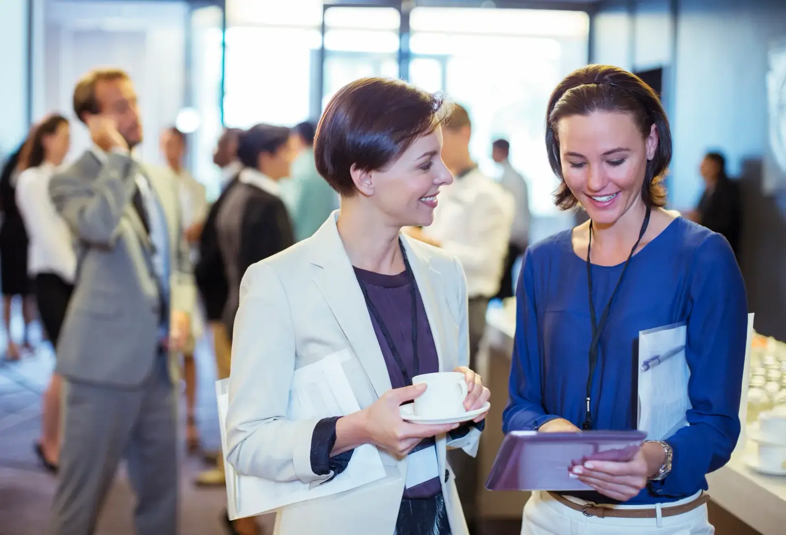 Two attendees with lanyards look at a tablet together during a busy event reception
