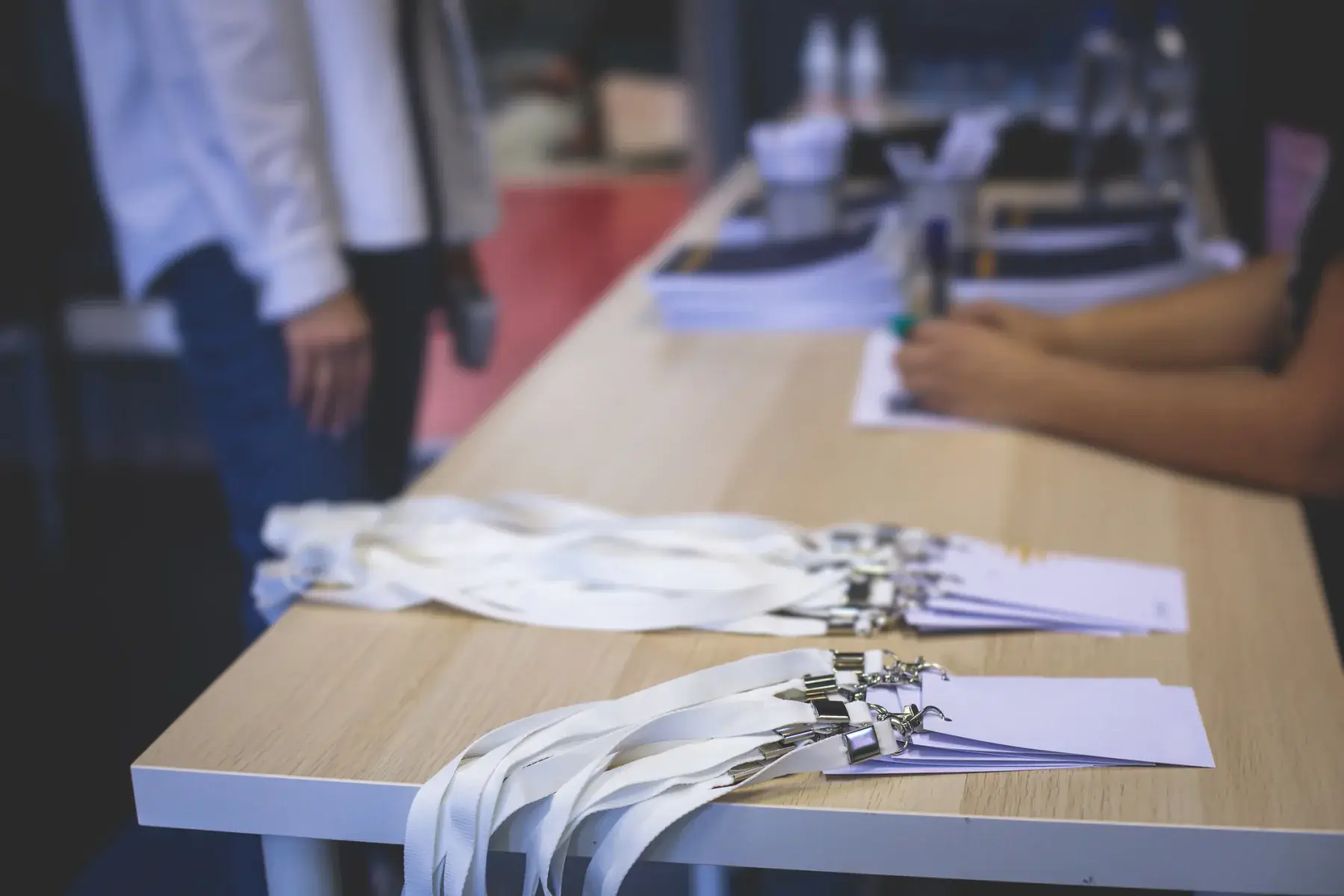 Blank badges on white lanyards lined up on a registration table, staff blurred behind